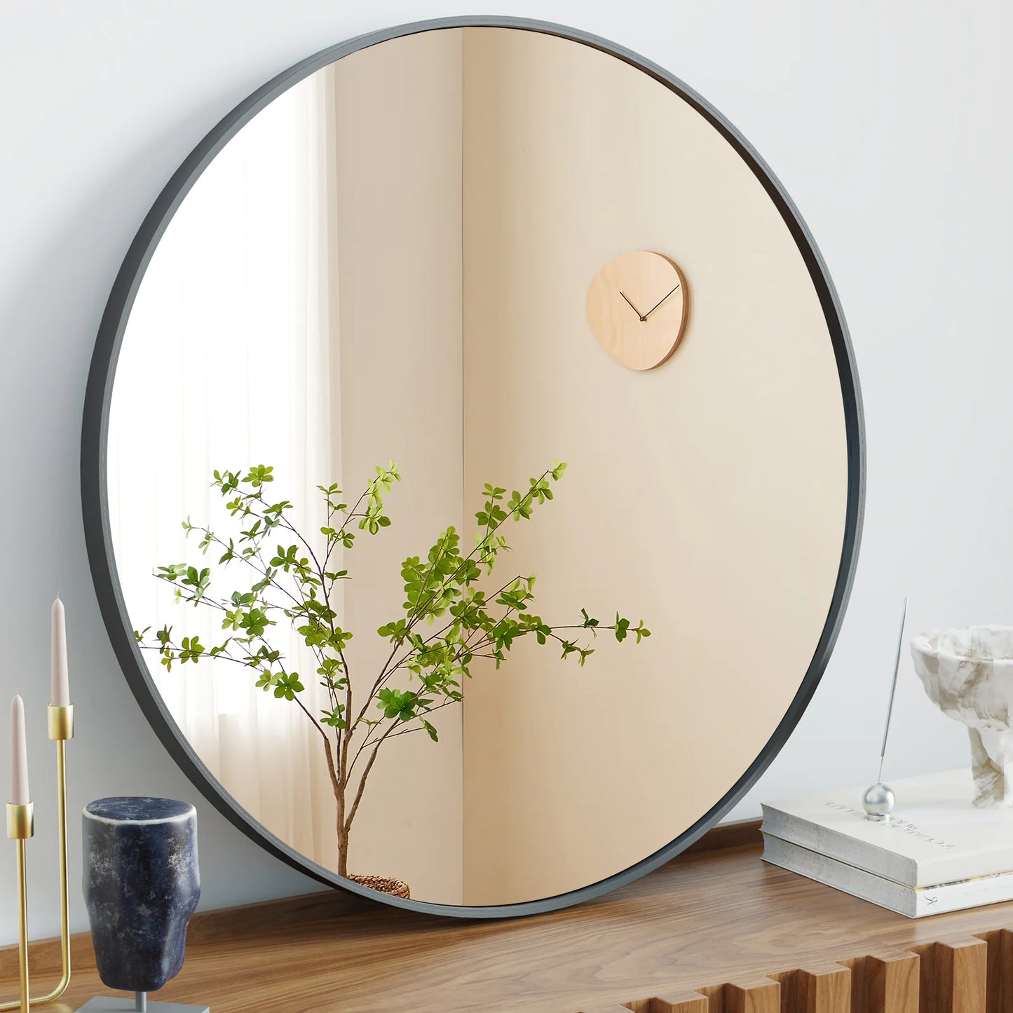 A black circular wall mirror hanging on a wall, reflecting a plant and a wooden clock.