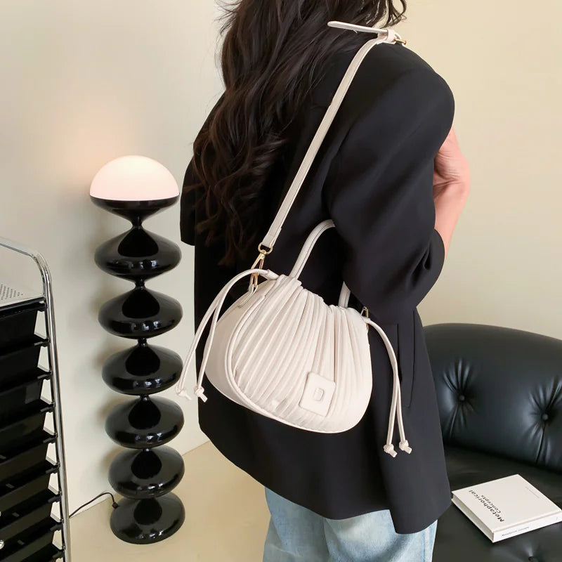 A person wearing a white dress and black vest holding a black PU leather bucket bag with a drawstring closure.