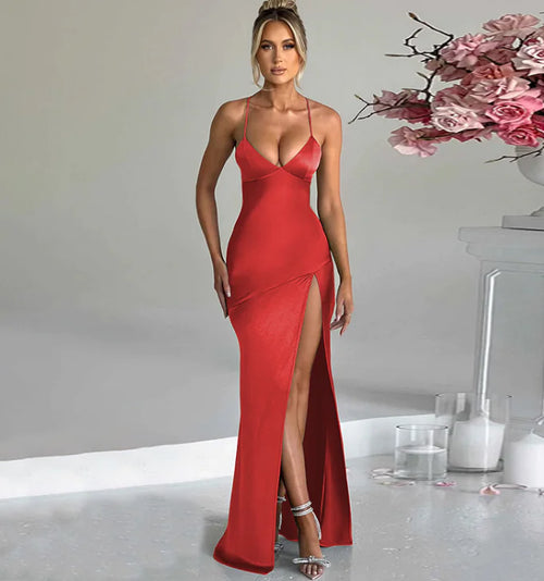 A woman in a red sleeveless maxi dress with a deep v-neckline and a high slit, standing in a room with a white floor and a floral arrangement in the background.