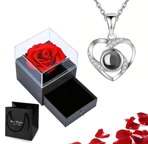 A silver-colored heart-shaped pendant necklace with a copper heart charm, displayed alongside a red rose inside a black and clear box, with scattered red petals around and a black packaging box.