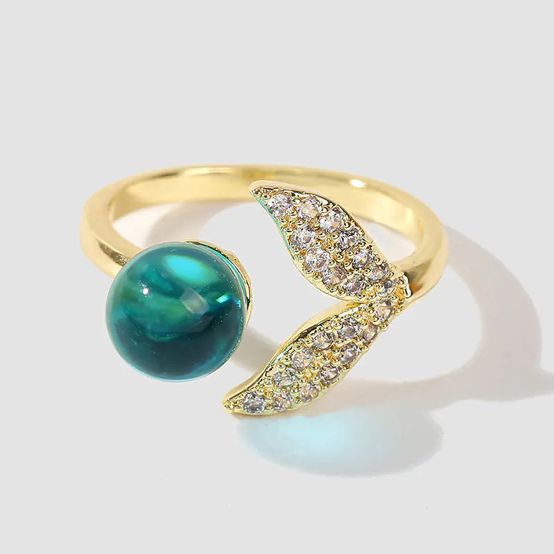 A gold-colored ring with a mermaid tail design adorned with small crystals, featuring a large green bead resembling an ocean creature.