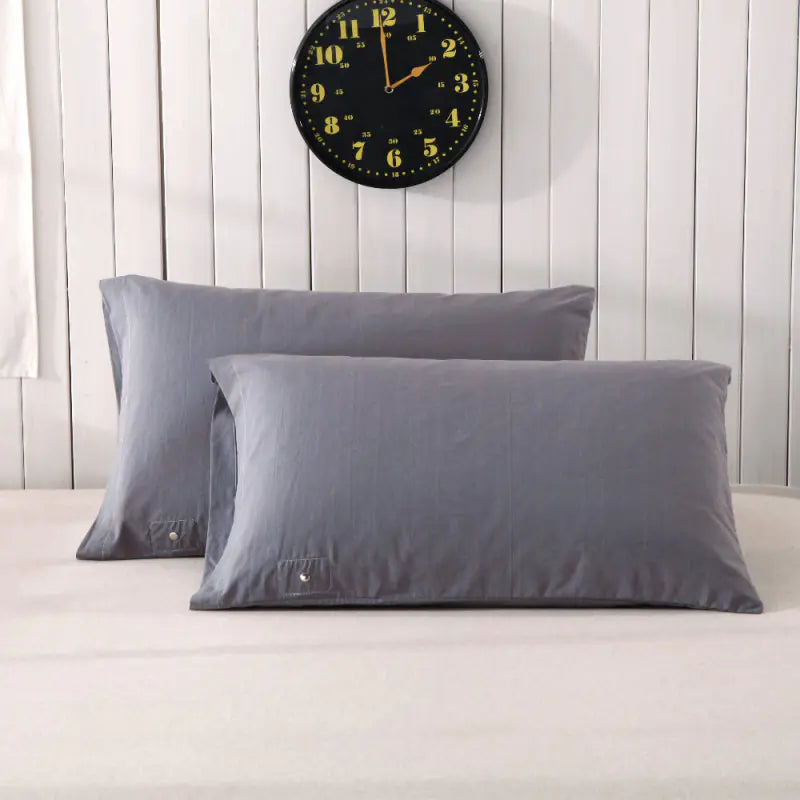 Two gray pillowcases on a bed with a white sheet, against a wooden wall with a clock above. The pillows are positioned at an angle, showcasing the brand tag on one of them.