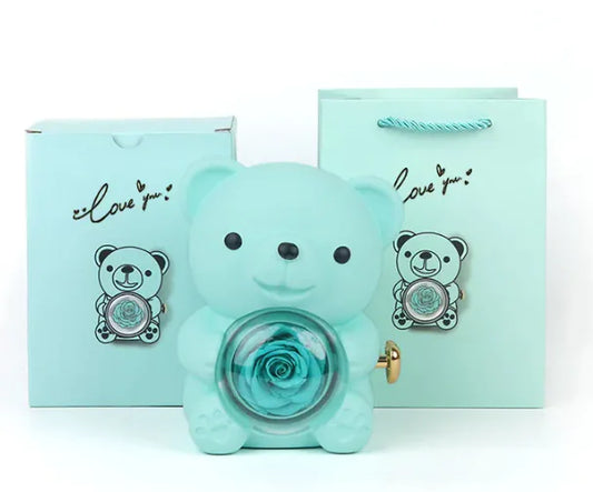 A teddy bear gift box in blue with a preserved rose and a necklace, alongside a rotating rose jewelry box, against a white background.