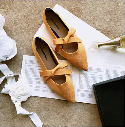 A pair of yellow flat casual boat shoes with bow Mary Jane style on a carpeted floor, surrounded by a white ribbon, a rose, and a pair of scissors.