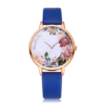 A floral pattern watch with a blue strap and gold-colored case, featuring a white dial adorned with a multicolored floral design.
