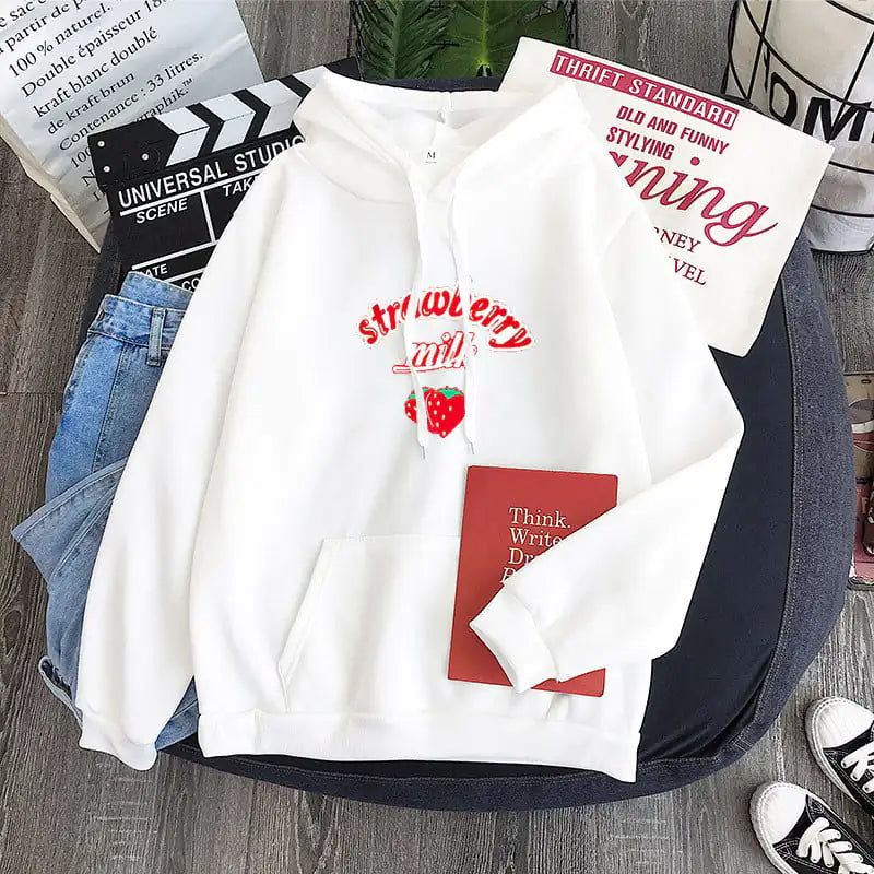 A black graphic sweatshirt with 'Strawberry Milk' text and a strawberry image on the front, displayed in a casual setting with jeans and sneakers.