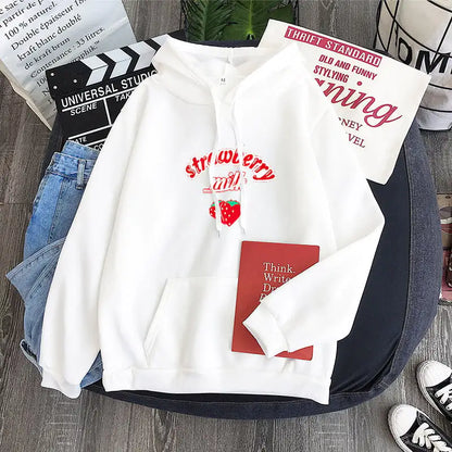 A black graphic sweatshirt with 'Strawberry Milk' text and a strawberry image on the front, displayed in a casual setting with jeans and sneakers.