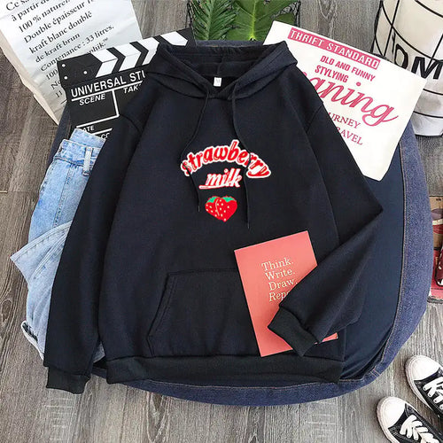 A black graphic sweatshirt with 'Strawberry Milk' text and a strawberry image on the front, displayed in a casual setting with jeans and sneakers.