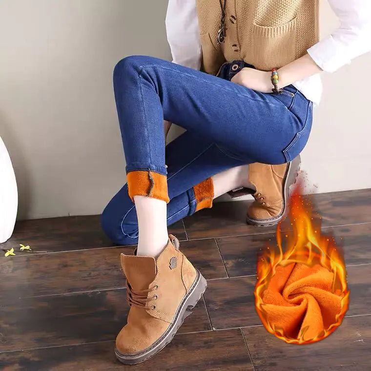 A pair of high-waisted plush jeans for women, in a dark color, styled with a pair of brown boots and an orange skirt, sitting against a wooden backdrop with a decorative item and a flame graphic effect on the image.
