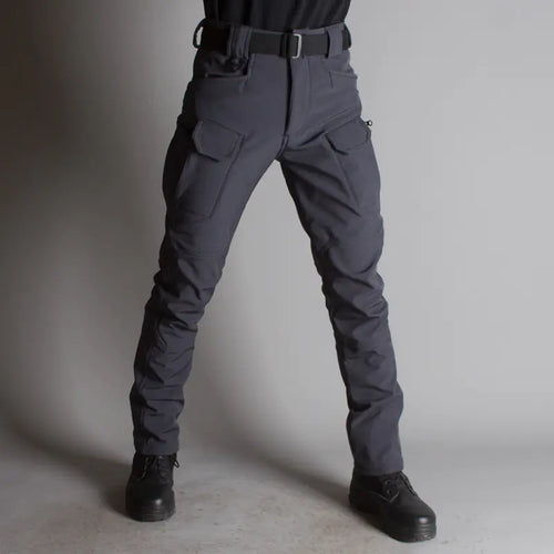 A person standing against a gray background, wearing black tactical pants with multiple pockets and a belt.