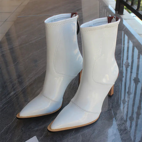 A pair of white leather ankle boots with pointed toes and high heels, featuring a back zipper closure, placed on a reflective surface.