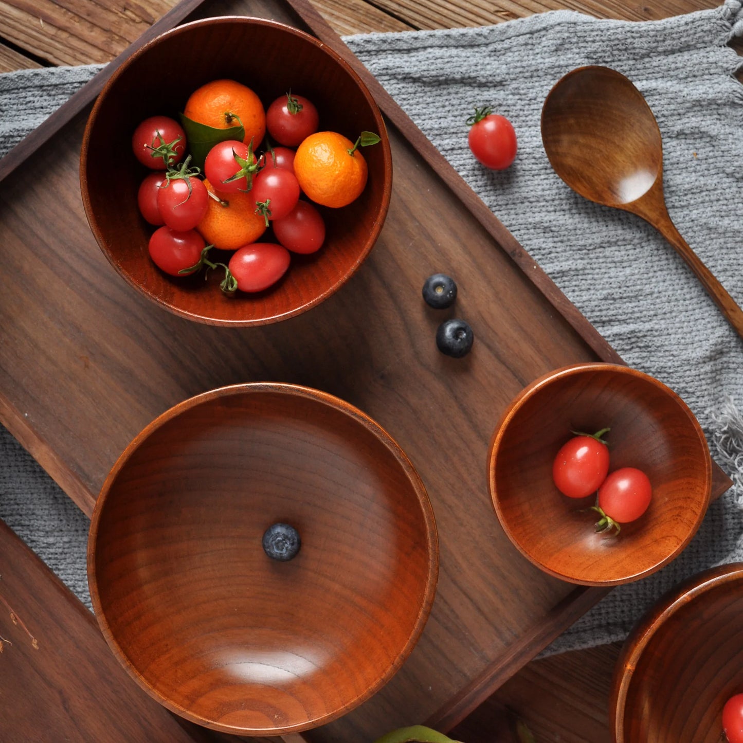 A set of Japanese wood bowls of various sizes arranged on a wooden tray, with one bowl containing colorful cherry tomatoes and another with a single blueberry. A wooden spoon and a grey cloth complete the setting.