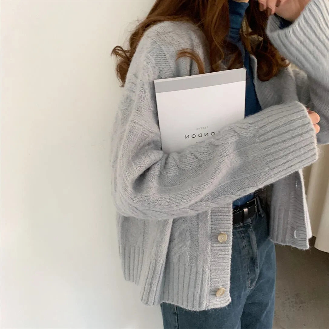A woman sitting and reading a book while wearing a beige knitted sweater cardigan jacket with long sleeves and a round neckline.