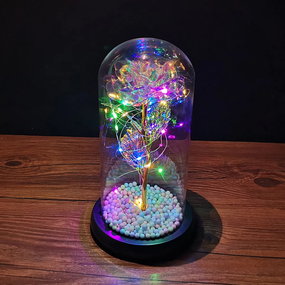 An LED Enchanted Glass Rose Decor with multicolored LED lights inside a clear glass dome, placed on a wooden stand with white pebble-like substances beneath.