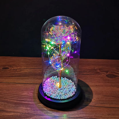 An LED Enchanted Glass Rose Decor with multicolored LED lights inside a clear glass dome, placed on a wooden stand with white pebble-like substances beneath.