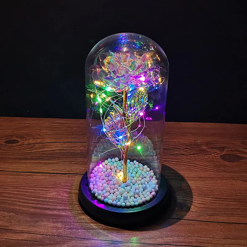 An LED Enchanted Glass Rose Decor with multicolored LED lights inside a clear glass dome, placed on a wooden stand with white pebble-like substances beneath.
