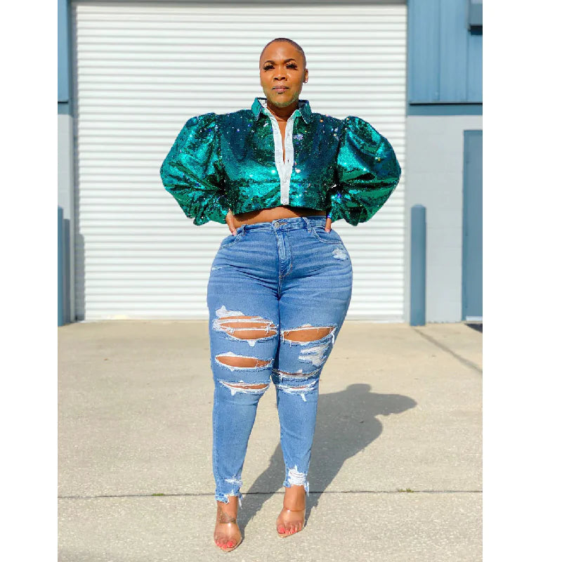 A woman standing in front of a blue door wearing large, ripped, light blue denim trousers and a puffed sleeve, multicolored top.