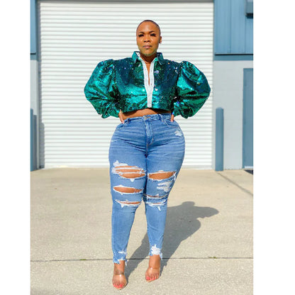 A woman standing in front of a blue door wearing large, ripped, light blue denim trousers and a puffed sleeve, multicolored top.