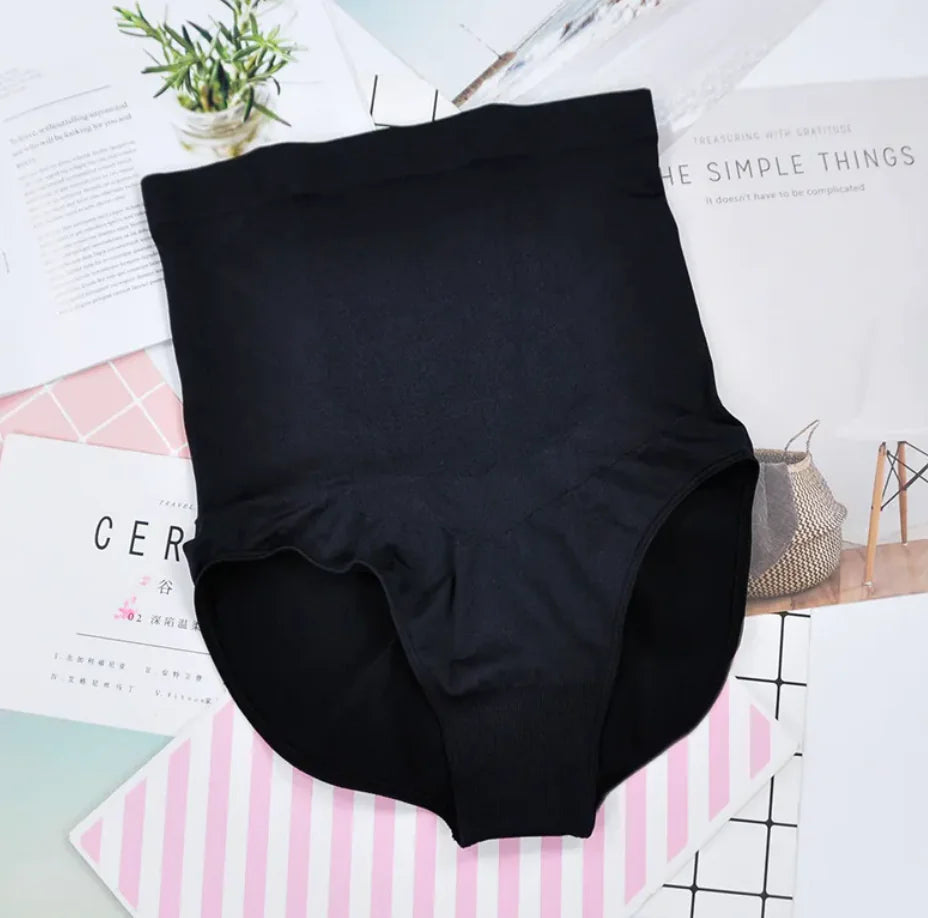 A pair of high-waisted black maternity panties displayed on a flat surface with a plant and magazine in the background.