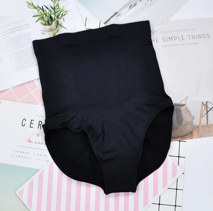 A pair of high-waisted black maternity panties displayed on a flat surface with a plant and magazine in the background.