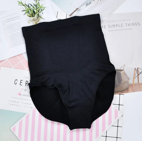 A pair of high-waisted black maternity panties displayed on a flat surface with a plant and magazine in the background.