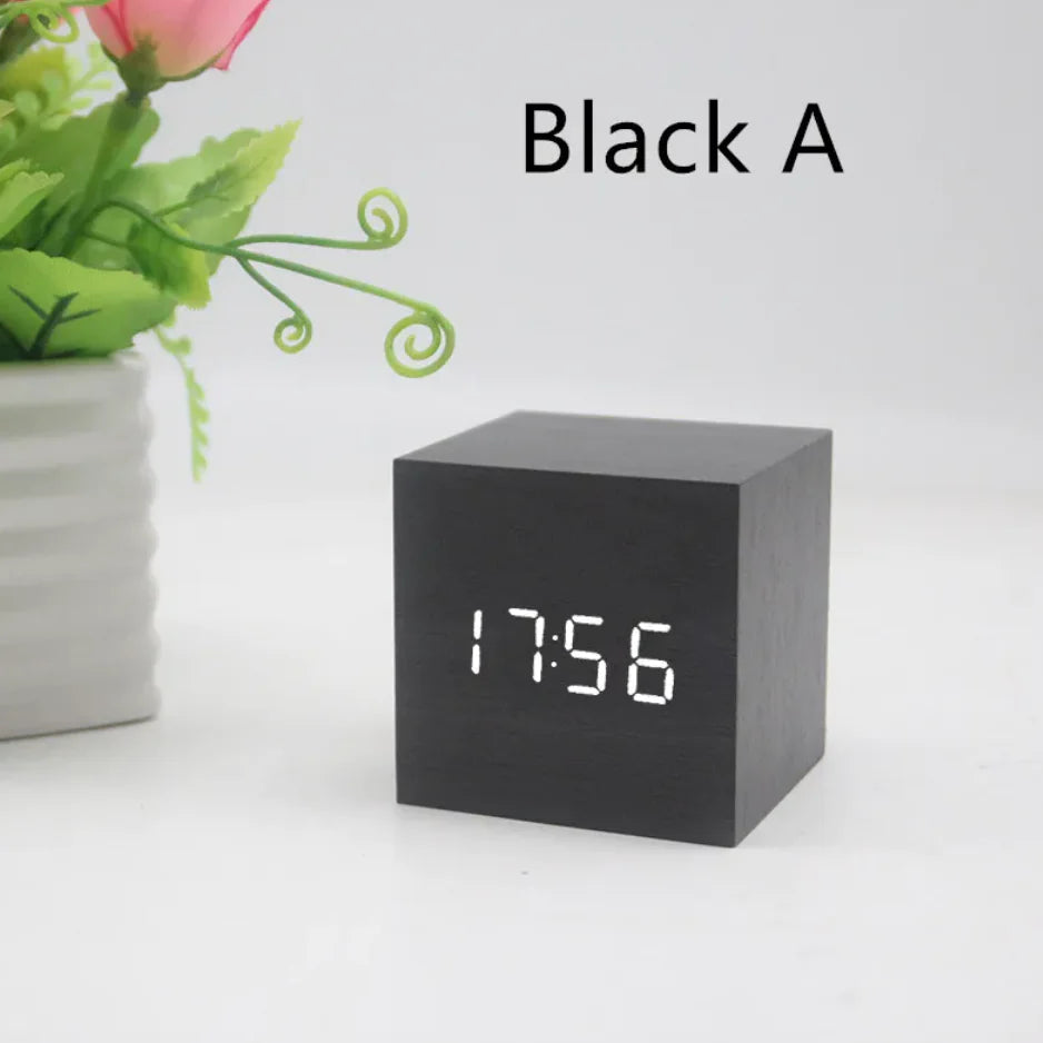 A black cube-shaped wooden digital clock displaying time on its LED interface, placed on a white surface next to a plant.