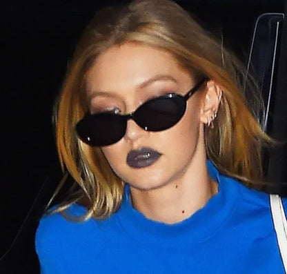 A woman with long blonde hair wears Retro Oval Sunglasses, dark grey lipstick, a blue top, and multiple small earrings, looking away from the camera against a dark background.