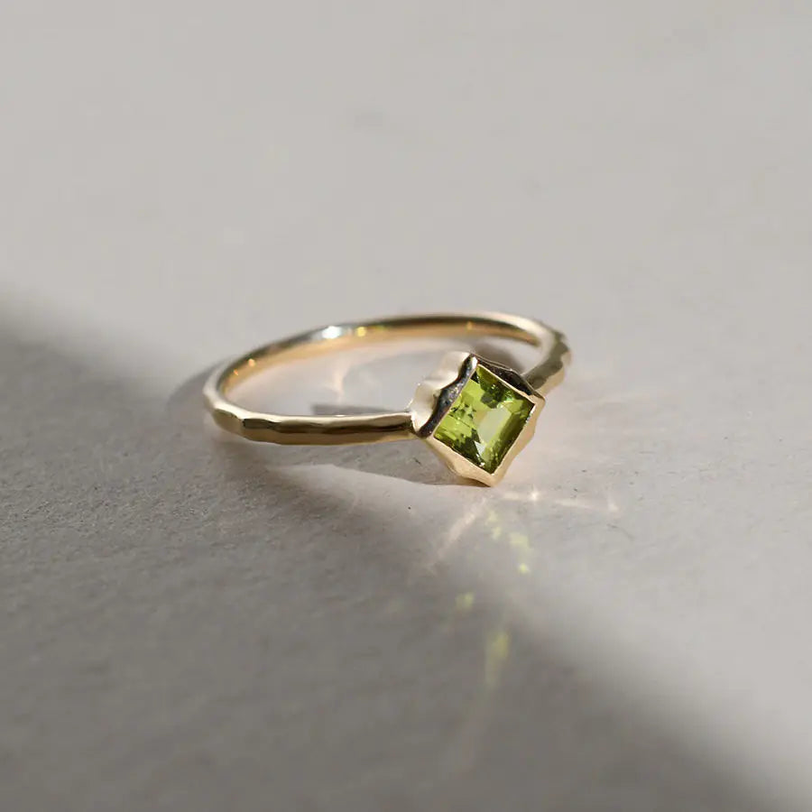 A silver ring with a square-cut green peridot stone, displayed against a neutral background with a source of light illuminating the stone.