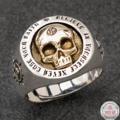 A silver ring with a central skull design, featuring intricate engravings and a small diamond-like detail on the top.