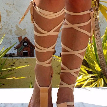 A pair of white thin strap flat sandals worn by a person standing against a stone wall background.