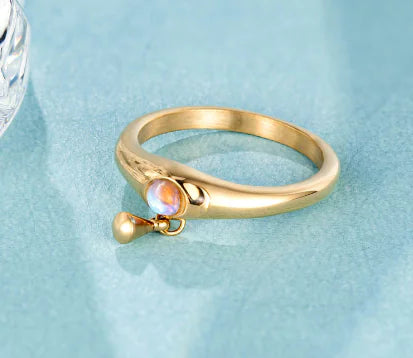 A gold-colored titanium steel ring with a pearl-like embellishment on a blue background.