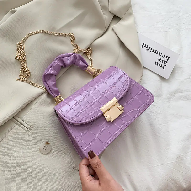 A purple crocodile pattern crossbody chain bag with a gold chain strap placed on a beige textile surface, with a part of a white garment and a text-bearing card visible in the background.