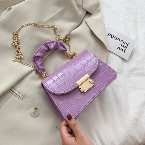 A purple crocodile pattern crossbody chain bag with a gold chain strap placed on a beige textile surface, with a part of a white garment and a text-bearing card visible in the background.