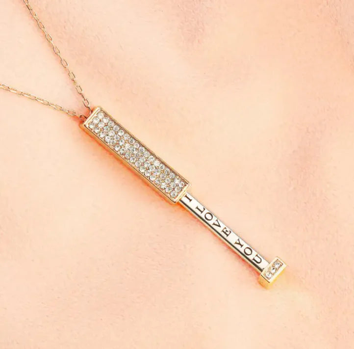 A gold-colored pendant necklace with a shimmering zircon stone inlaid into a bar-shaped design, displayed against a pink background.
