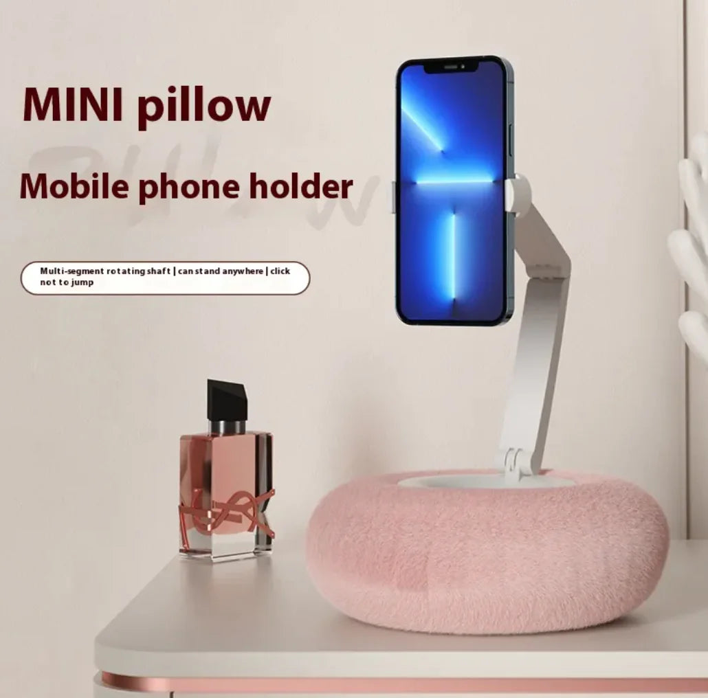 A plush pillow stand in pink color, advertised as a mobile phone holder, placed on a flat surface with a mobile phone upright on it, and a perfume bottle in the background.