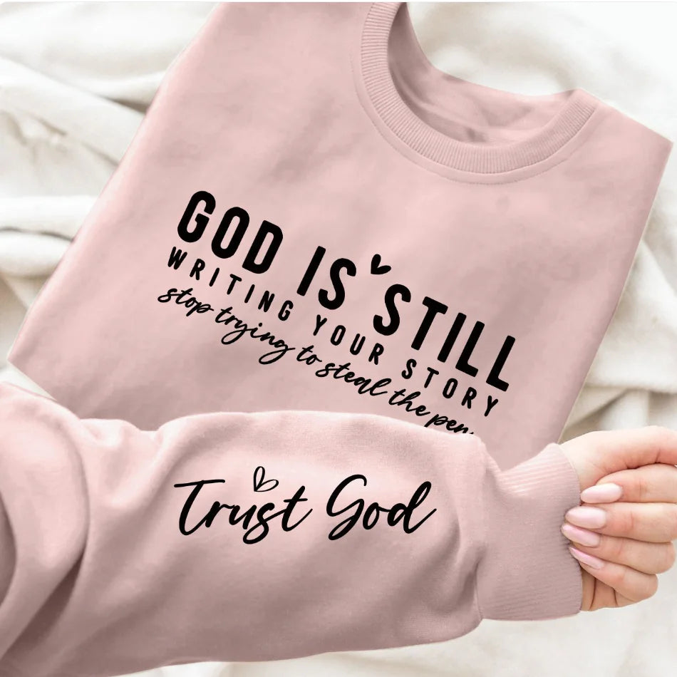 A gray crew neck sweatshirt with a text design 'GOD IS STILL WRITING YOUR STORY' printed on the front, paired with blue denim jeans.