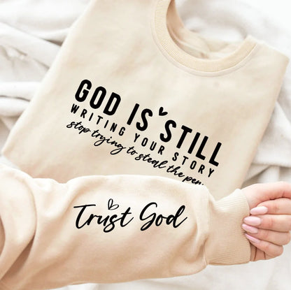 A gray crew neck sweatshirt with a text design 'GOD IS STILL WRITING YOUR STORY' printed on the front, paired with blue denim jeans.