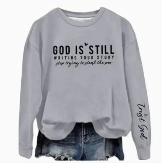 A gray crew neck sweatshirt with a text design 'GOD IS STILL WRITING YOUR STORY' printed on the front, paired with blue denim jeans.