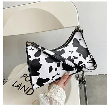 A black and white cow print baguette bag with a woman's hand holding it, displayed in front of a white background with the text 'POE LAND'.
