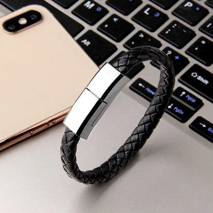 A bracelet charging cable designed to be worn on the wrist, displayed on a laptop next to a smartphone, showcasing its brown leather strap and gold-colored clasp.