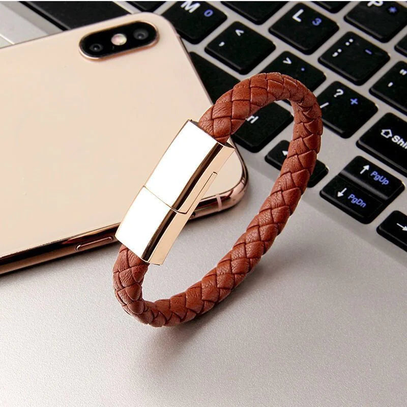 A bracelet charging cable designed to be worn on the wrist, displayed on a laptop next to a smartphone, showcasing its brown leather strap and gold-colored clasp.